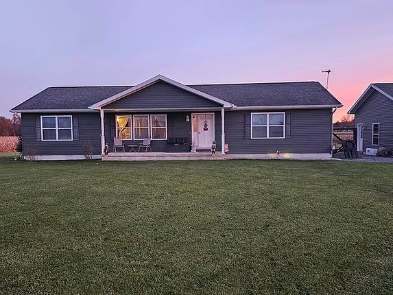 A gray, single-story ranch-style home with a front porch, set against a sunset sky with a large lawn in the foreground.