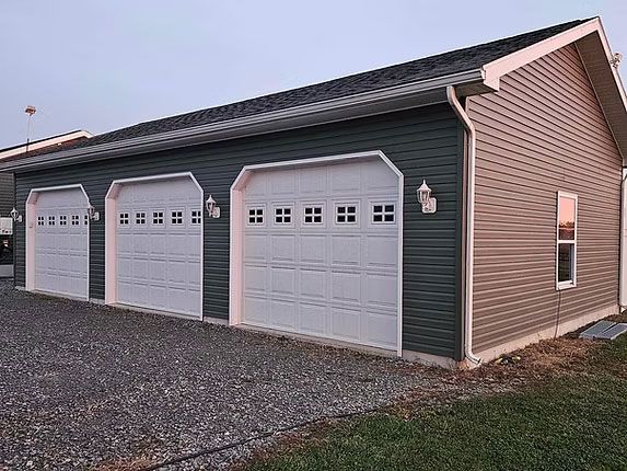 A three-car garage with white doors, dark gray siding on the front, tan siding on the side, and a gravel driveway.