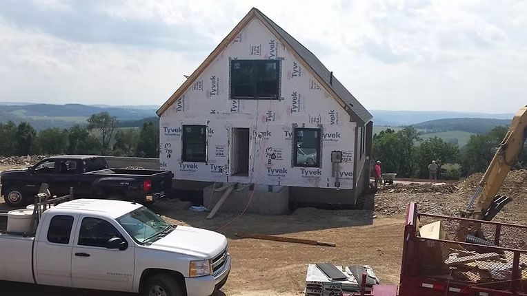 A house under construction with white Tyvek siding sits on a hilltop, with work trucks and equipment parked nearby.