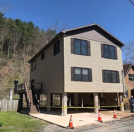 A tan two-story house on stilts with a side deck and stairs, surrounded by yellow caution tape.