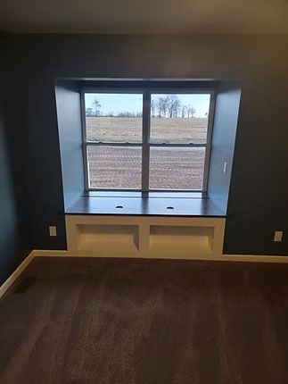 A built-in window seat with storage cubbies underneath, centered in a room with dark blue walls and brown carpeting.