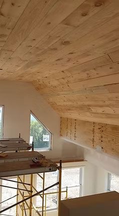 An unfinished attic room featuring a sloped wooden ceiling, white walls, angled windows, and construction scaffolding.