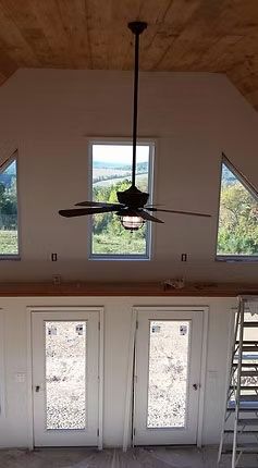 A vaulted room features a dark ceiling fan, three triangular windows, two glass-paneled doors, and a metal ladder.