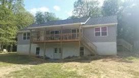 A two-story house with light gray siding and a white painted foundation, featuring a large wooden deck and back stairs.