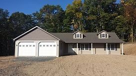 A single-story, light-tan house with a two-car garage, a covered front porch, two dormer windows, and a brown roof.