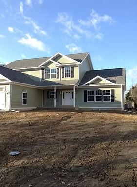 A newly built light-green two-story suburban house with a gray shingled roof and an unfinished dirt yard under a blue sky.