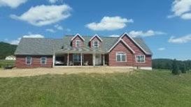 A red brick house with a covered porch and dormer windows, situated in a green field under a blue sky.