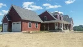 A two-story red farmhouse with a wrap-around porch and an attached two-car garage sits on an open, dirt-covered lot.
