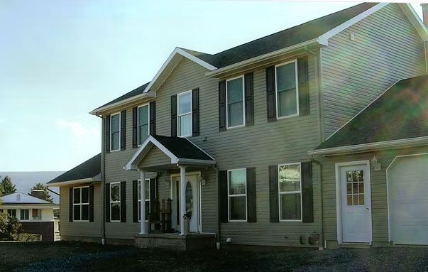 A two-story, light-colored vinyl-sided suburban home with dark shutters and a small front porch under a sunny blue sky.