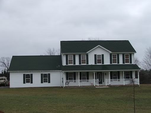 A white, two-story house with a green roof, dark shutters, and a front porch, set on a grassy lawn under a cloudy sky.