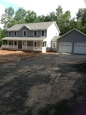 A two-story grey farmhouse with a wraparound porch and an attached two-car garage, set against a background of trees.