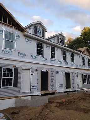 A multi-unit building under construction, featuring white Tyvek wrap, installed windows, and two roof dormers.