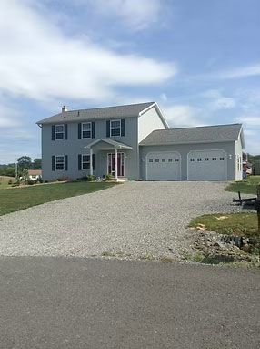 A two-story grey house with a white front porch and an attached two-car garage, viewed from a gravel driveway.