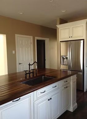 A kitchen island with a wood countertop and dark sink, featuring white cabinets and a stainless steel refrigerator.
