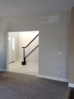 An interior view from a carpeted room into an entryway with wood flooring, a staircase with black railing, and a door.
