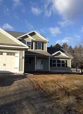 A two-story green house with a white garage and door, under a blue sky with bare trees and unfinished dirt in the yard.