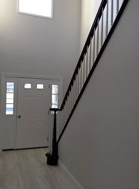 A bright foyer featuring light wood floors, a white front door with windows, and a staircase with a dark wood railing.
