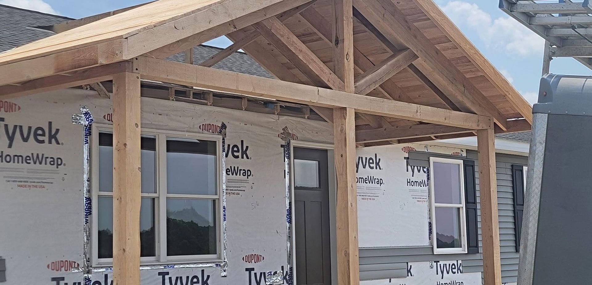 A house under construction with exposed wooden beams, a framed porch roof, and walls covered in white Tyvek wrap.