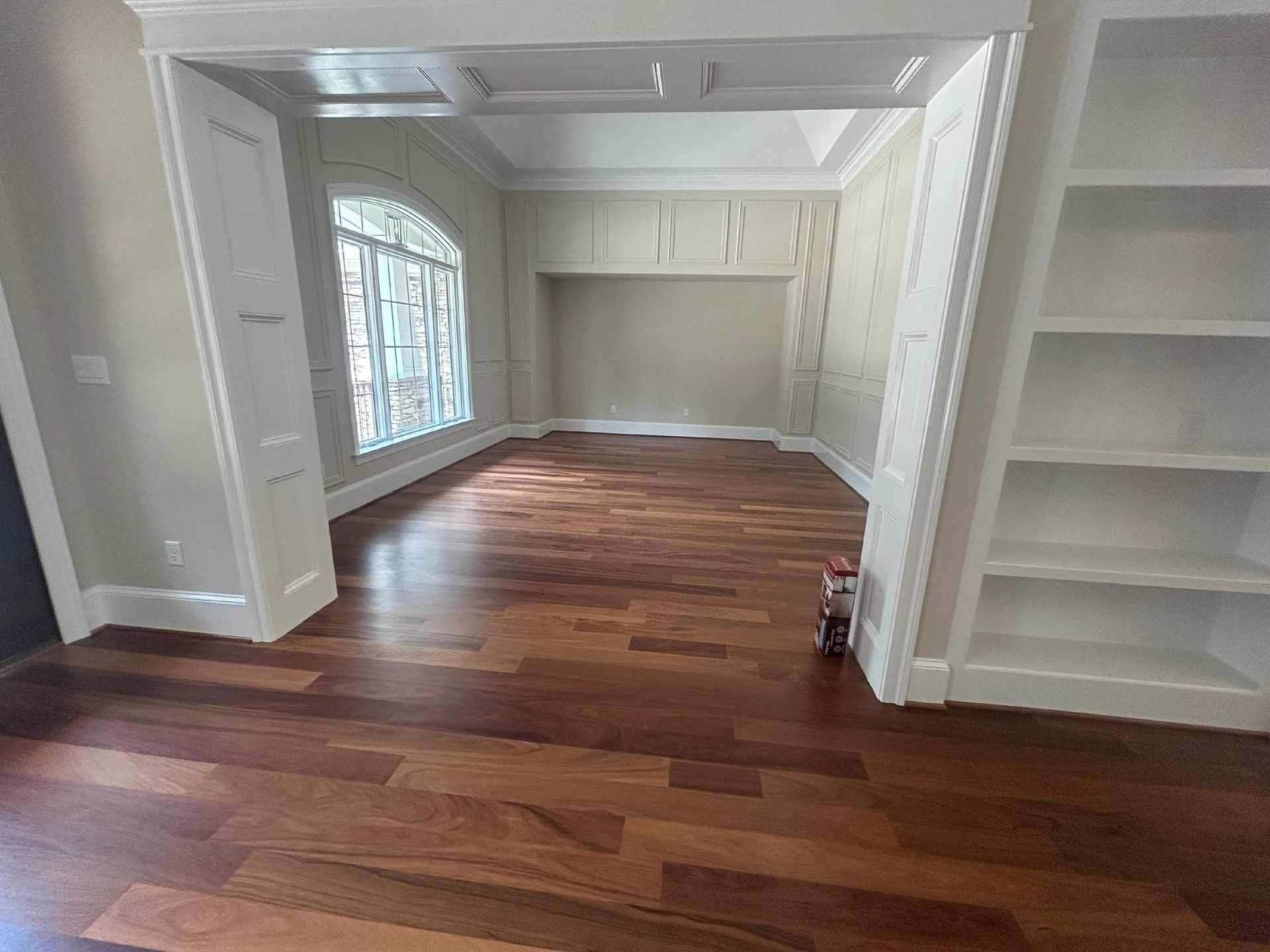 A spacious room with polished dark wood floors, white built-in shelving, and a large window looking onto a bright area.