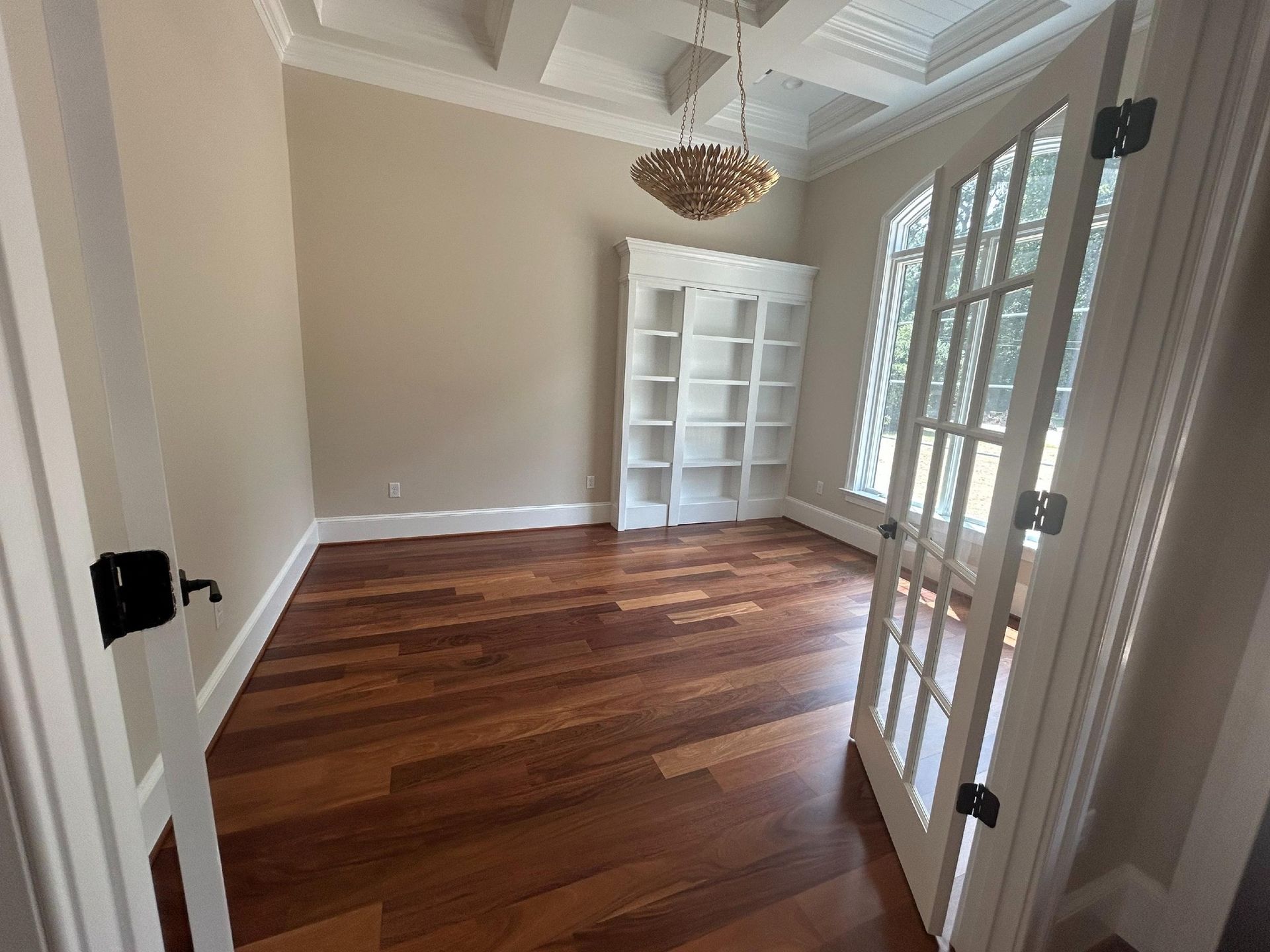 A room with hardwood floors, a white bookshelf, beige walls, and a coffered ceiling with a chandelier viewed through doors.