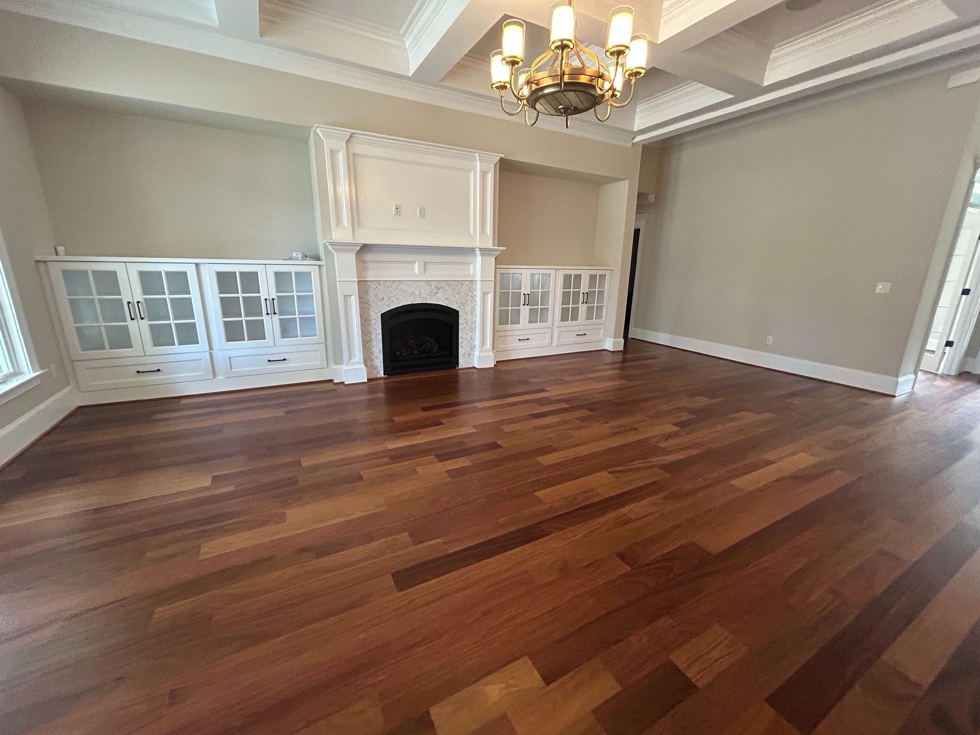 Spacious living room with rich hardwood floors, built-in white cabinets, a fireplace, and a coffered ceiling with chandelier.