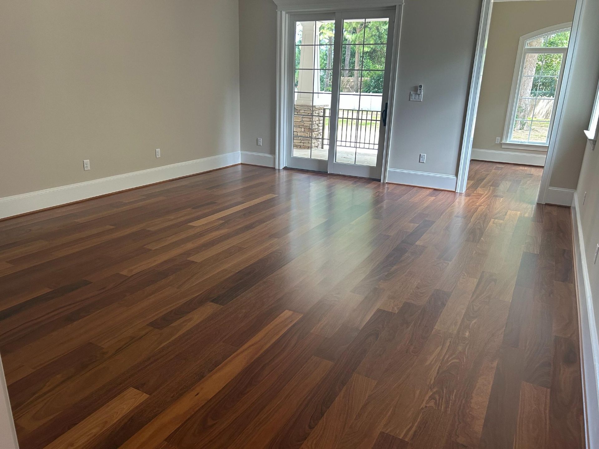 An empty room with rich, warm-toned wood flooring, white trim, and a glass sliding door leading to an outdoor deck.