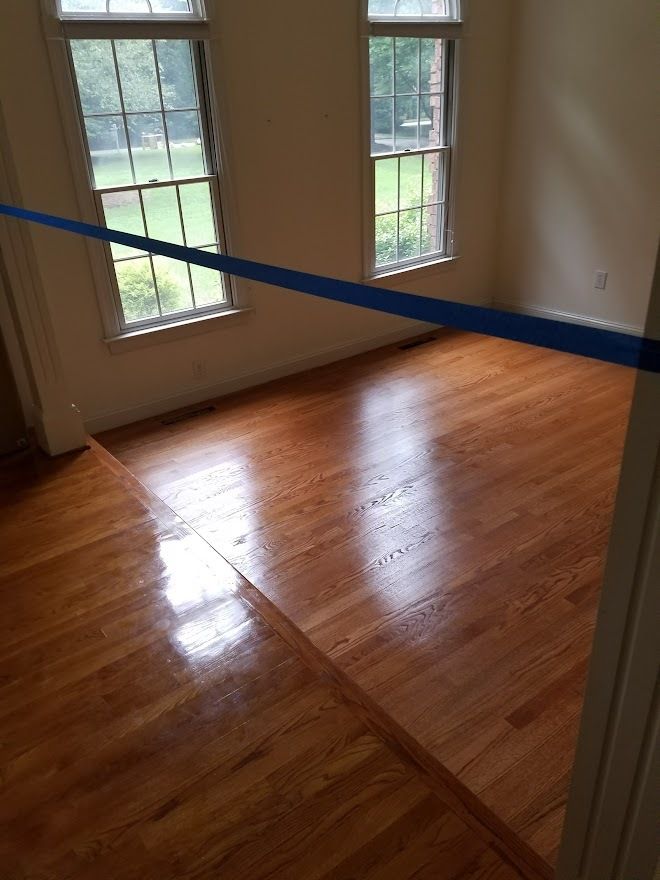 An empty room with polished hardwood floors and two windows, viewed through a doorway with a horizontal blue tape line.