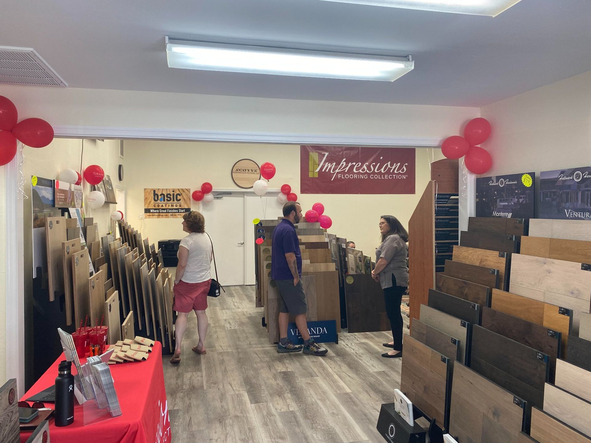 Inside a flooring showroom with wood sample displays, two people stand and talk near the back, with red balloons nearby.