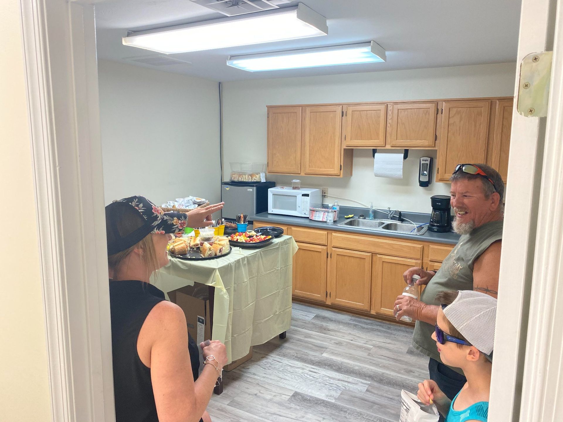 A group of people standing in a kitchen, looking toward a table with snacks, viewed through a doorway.