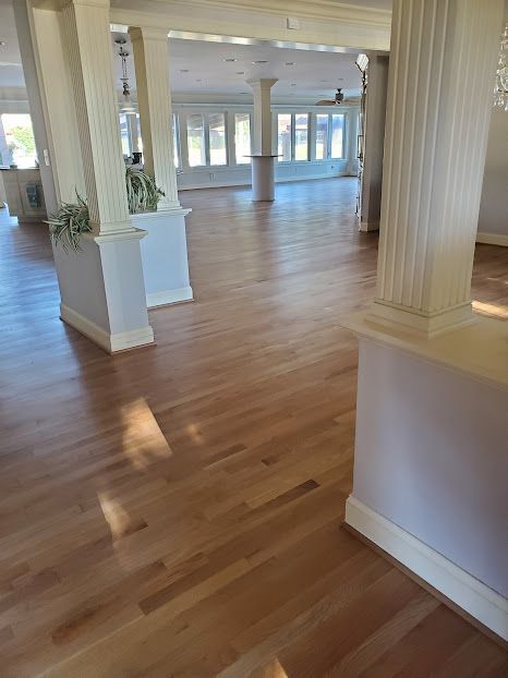 Interior view of a spacious room with hardwood floors, white columns, and large windows bringing in natural light.
