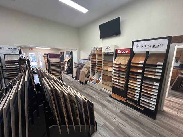 An interior view of a flooring showroom featuring rows of wood, tile, and vinyl sample displays under recessed lighting.