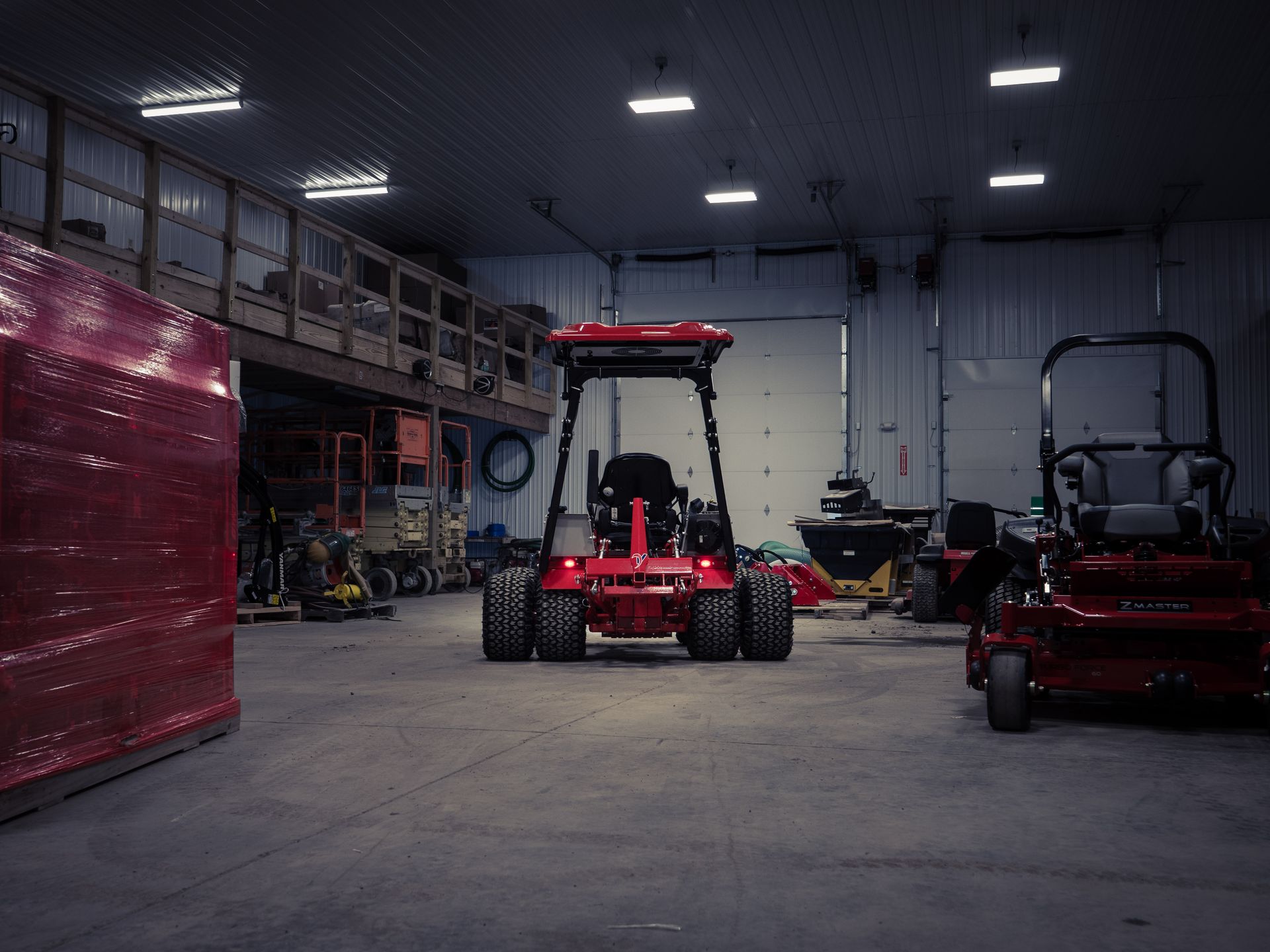 A red tractor is parked in a warehouse next to a lawn mower.