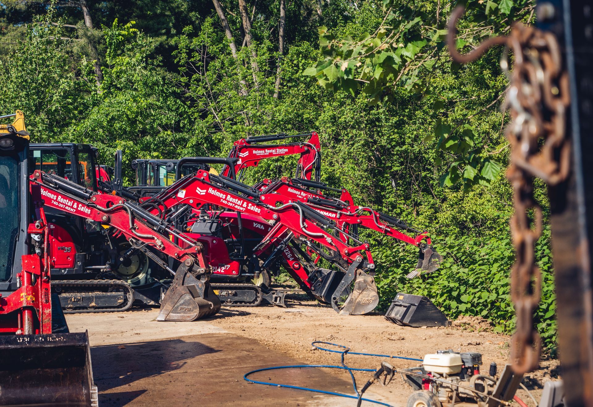 A row of red excavators are parked in a dirt lot.