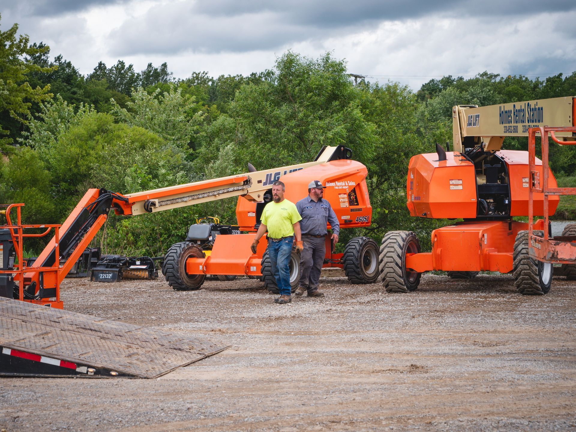 Two men are standing next to a jlg lift in a gravel lot.