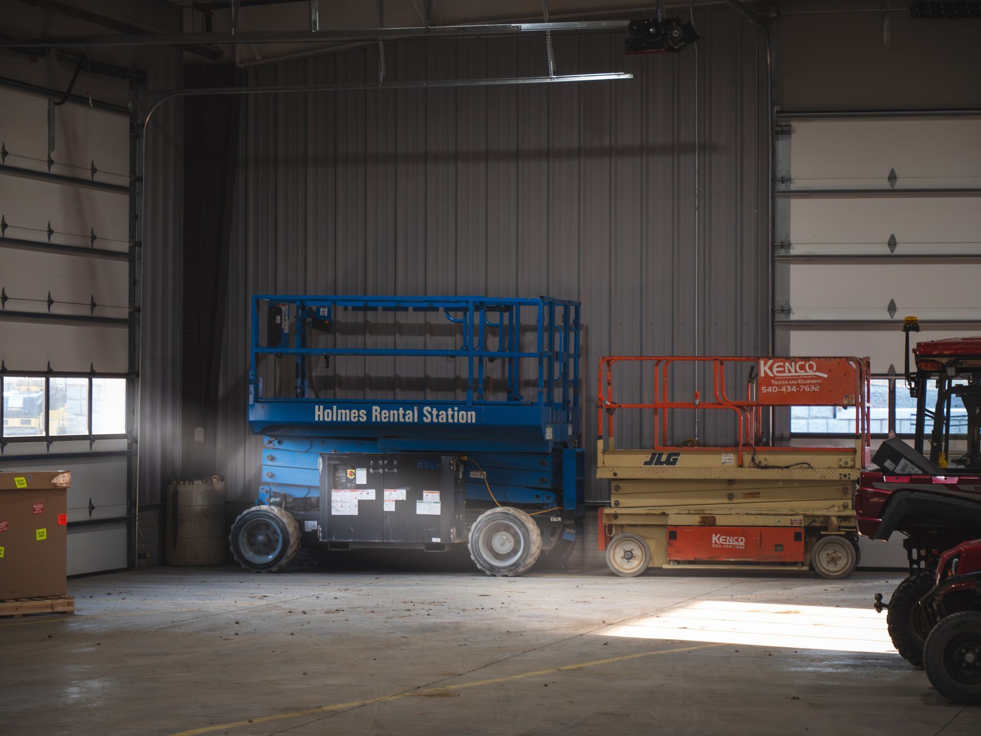 Two scissor lifts are parked in a garage next to a tractor.