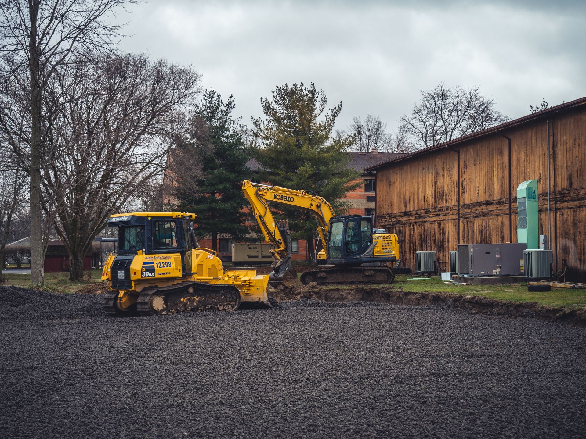 Two construction vehicles are parked in a gravel lot in front of a wooden building.