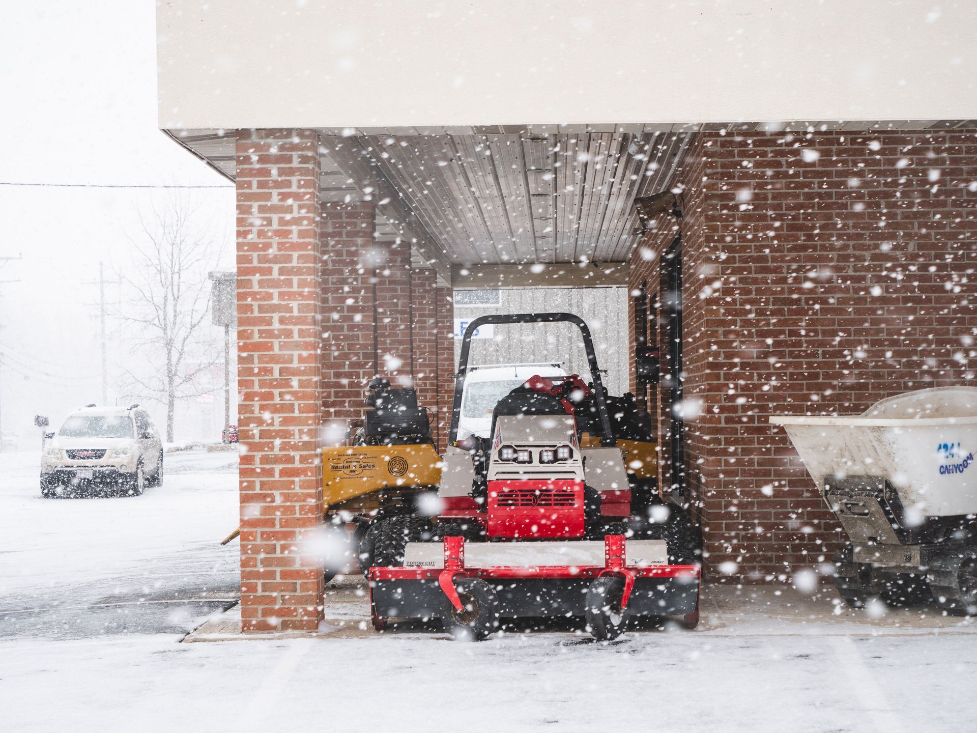 A snow blower is parked under a brick building in the snow.