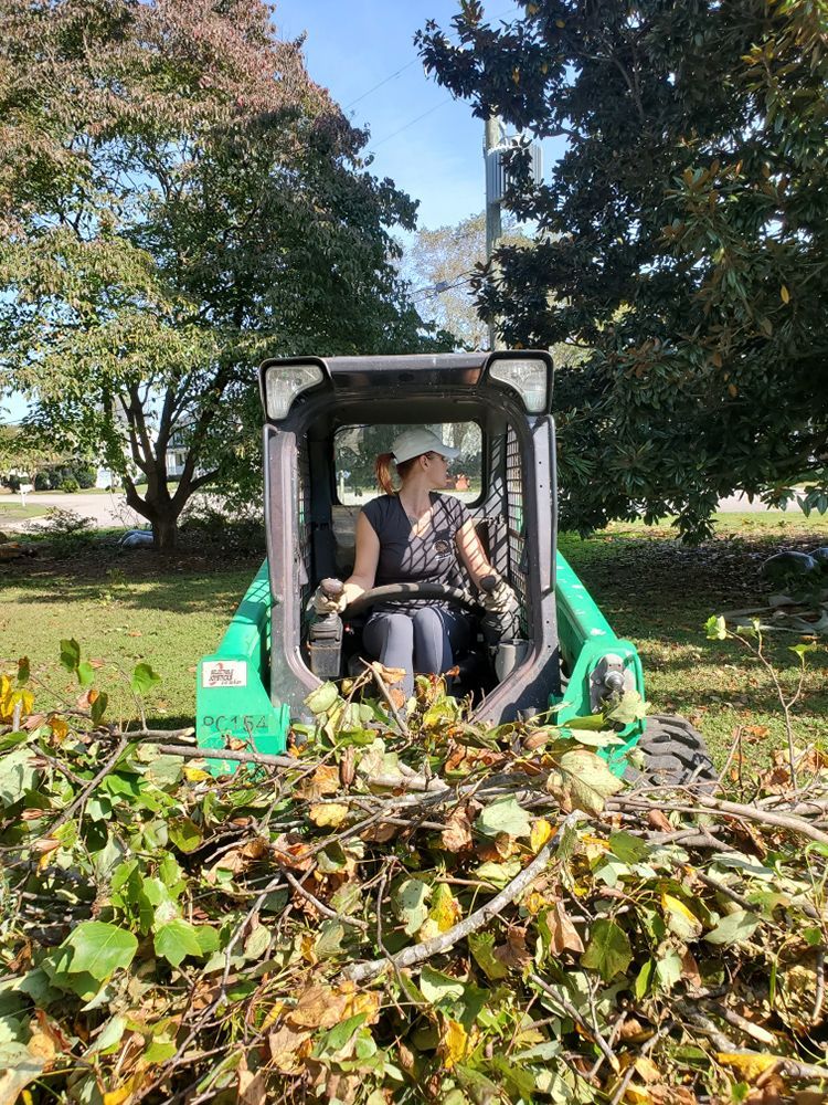 A woman is driving a green tractor through a pile of branches.