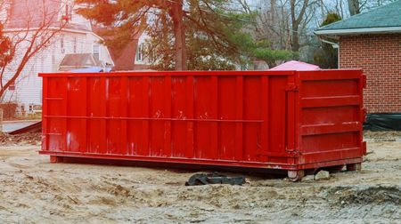 Red dumpster on a construction site; houses and trees in the background.
