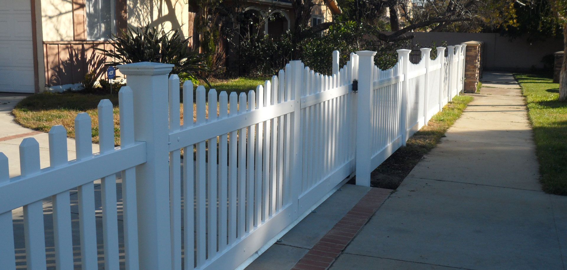 White picket fence along a sunny sidewalk beside a house and yard