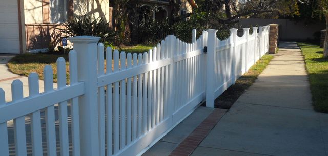 White picket fence along a sunny sidewalk beside a house and yard