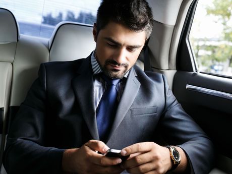 A man in a suit and tie is sitting in the back seat of a car looking at his cell phone.