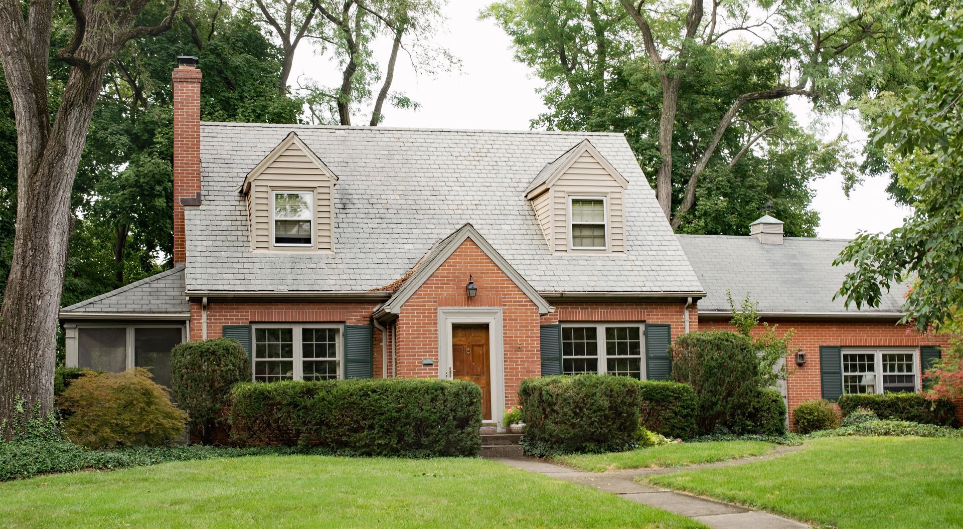 Brick house with a gabled roof, chimney, and front walkway, surrounded by green lawn and trees