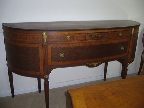 Curved wooden buffet with drawers, brass hardware, and slender legs against a white wall.