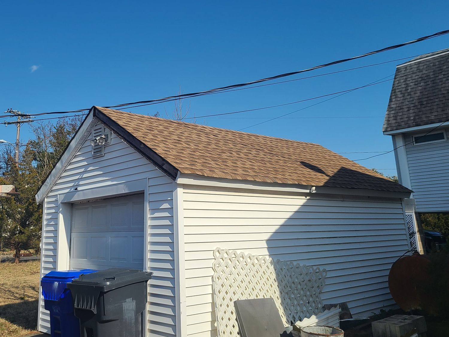 A white garage with a brown roof and a blue trash can in front of it.