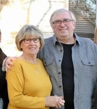 Smiling couple standing outdoors, woman in yellow sweater and man in gray shirt with arm around her.