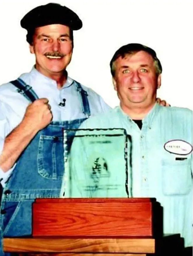 Two men in work aprons stand behind a wooden box, smiling at the camera.