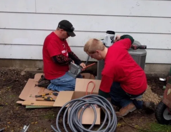 Two workers in red shirts installing equipment outdoors beside coiled cables and tools.