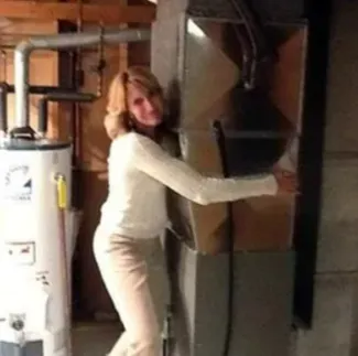 Woman posing beside a basement furnace and ductwork, hugging a metal vent with a slight smile.