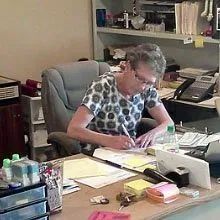 Person writing at a cluttered office desk with papers, phone, and supplies.
